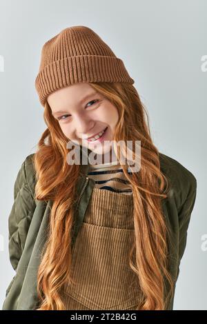 bambina elegante, ragazza allegra con cappello a maglia e capispalla che guarda la fotocamera su sfondo grigio Foto Stock