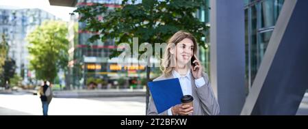 Occupato donna d'affari beve caffè, parla sul telefono cellulare, tiene documenti per il lavoro, passeggiate nel centro della città vicino ufficio Foto Stock