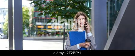 Occupato donna d'affari beve caffè, parla sul telefono cellulare, tiene documenti per il lavoro, passeggiate nel centro della città vicino ufficio Foto Stock