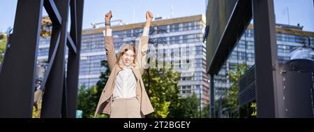 Ritratto di felice giovane donna d'affari alzare le mani, pugno pompa, celebra la vittoria, vince sth, raggiunge obiettivo, si trova all'aperto sulla strada Foto Stock