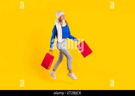 Grandi vendite stagionali. Lady Shoopaholic che tiene le borse della spesa e corre a mezz'aria, indossando maglione, sciarpa e cappello Foto Stock