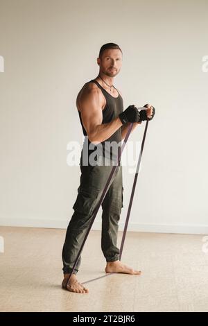 Un uomo che indossa abbigliamento sportivo nero è impegnato nel fitness della forza utilizzando un anello in gomma in interni. Foto Stock