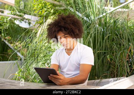 Concentrato bellissimo giovane etnico, vestito con abiti casual, lavora e disegna su un tablet all'aperto, in un'area verde nella natura. Spazio verde aperto Foto Stock