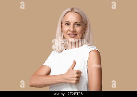 Donna matura con cerotto medico sul braccio che mostra pollice-up su sfondo beige. Concetto di vaccinazione Foto Stock