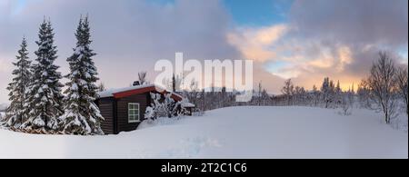 Un piccolo rifugio di caccia innevato sulle montagne norvegesi con tracce di animali in primo piano e vette di montagna sullo sfondo Foto Stock