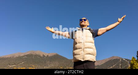 Banner bello 40 anni maschio Tourist con Open Arms, Mountains, Blue Sky sullo sfondo. Escursionista maschile con zaino fa trekking, trekking nella natura autunnale Foto Stock