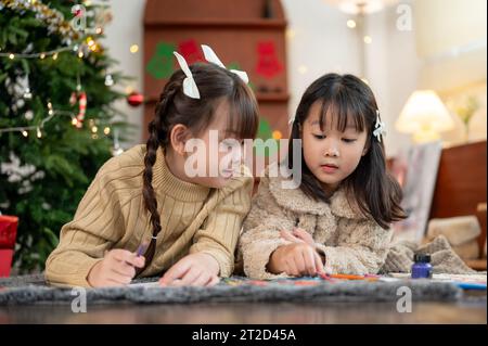 Due giovani ragazze asiatiche carine e felici sono sdraiate sul pavimento del soggiorno vicino all'albero di Natale e si divertono a disegnare e colorare insieme. Kid Leis Foto Stock