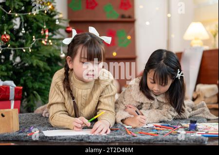 Due giovani ragazze asiatiche carine e felici sono sdraiate sul pavimento del soggiorno vicino all'albero di Natale e si divertono a disegnare e dipingere insieme. Kid Leis Foto Stock