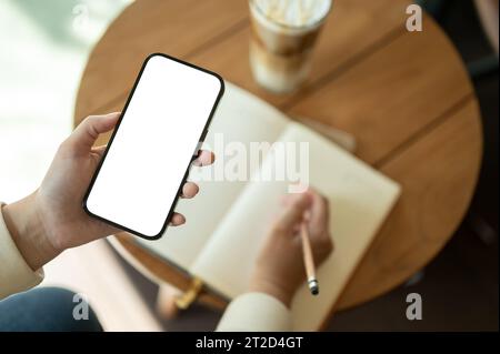Immagine dall'alto di una donna che usa lo smartphone mentre tiene il suo diario o scrive qualcosa nel suo libro in un bar. Uno smartphone con schermo bianco mo Foto Stock