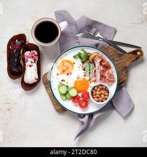 Piatto tradizionale inglese per la colazione con strisce di pancetta, uova soleggiate, verdure e torta su sfondo chiaro. Foto Stock