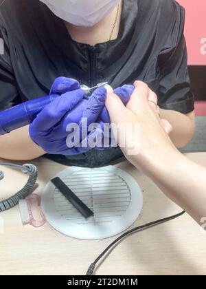 La donna, la ragazza il padrone sulla manicure, un servizio del chiodo fa una manicure in guanti blu scuro sulle mani il dispositivo una lima per i chiodi. Foto Stock