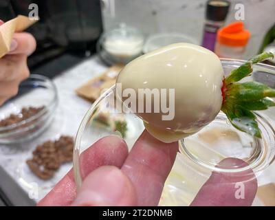 Fragola dolce a strati rossa temprata in un bel cioccolato belga fuso bianco con decorazioni su un bastoncino. Produzione di fragole ricoperte di cioccolato. Foto Stock