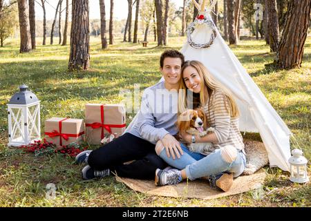 Famiglia sorridente di una giovane coppia e un cane che posa con decorazioni natalizie invernali nella foresta Foto Stock