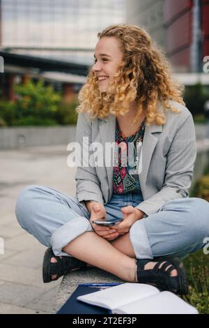 Ritratto all'aperto di una studentessa sorridente con smartphone e libri di lavoro seduti all'esterno del campus. Riposa dopo le lezioni. Foto Stock