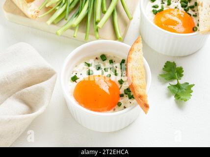 Uova cotte al forno (uova in cocotte) con panna, сheese e cipolla verde in ramoscelli su sfondo bianco. Servito con pane tostato caldo per condire. Vista ravvicinata Foto Stock