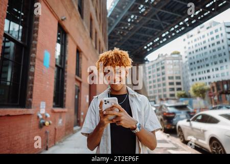 Un giovane ragazzo affascinante cammina dall'altra parte della strada, scrivendo messaggi su uno smartphone e sorridendo Foto Stock