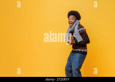 Felice giovane afro-americano con maglione e sciarpa natalizi che esprime una sensazione di sorpresa su uno sfondo giallo isolato con la foto in studio dello spazio di copia Foto Stock