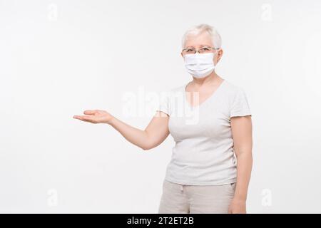 Ritratto di anziana donna caucasica che indossa la maschera medica con gester a mano aperta per copiare spazio in sfondo bianco isolato studio girato, auto di salute Foto Stock