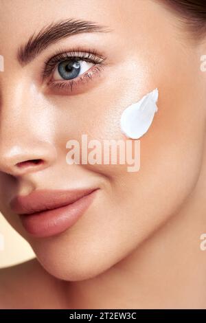 Bella donna che applica crema idratante sul viso. Foto di una donna sorridente con trucco perfetto su sfondo beige. Concetto di bellezza Foto Stock