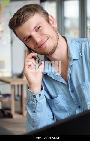 Bell'uomo parla al telefono e sorride. L'operatore risponde alla chiamata telefonica. Il ragazzo caucasico in camicia di denim è seduto in ufficio e parla al telefono cellulare. Emozioni positive. Servizio di supporto cordiale. Foto Stock