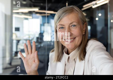 Foto ravvicinata di un'anziana donna d'affari sorridente che parla con una videocamera in ufficio, saltando la mano e salutando. Foto Stock