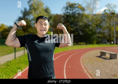 Ritratto di uno sportivo asiatico in piedi in uno stadio e che mostra alla fotocamera un gesto di potenza con le mani, mostrando i muscoli tonici e il corpo. Foto Stock