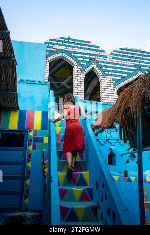 Una giovane donna con un vestito rosso che sale alcune scalinate colorate che salgono su una splendida terrazza di una tradizionale casa blu in un villaggio nubiano vicino Foto Stock