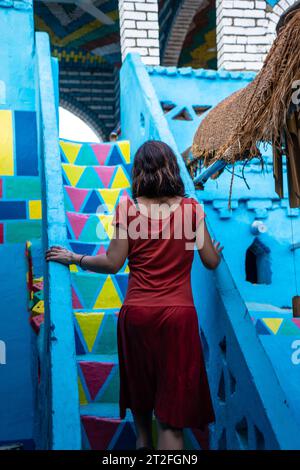 Una giovane donna con un vestito rosso che sale alcune scalinate colorate che salgono su una splendida terrazza di una tradizionale casa blu in un villaggio nubiano vicino Foto Stock