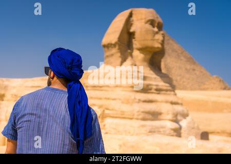 Un giovane turista che gode e ammira la grande Sfinge di Giza vestita di blu e di un turbante blu. Cairo, Egitto, Africa Foto Stock