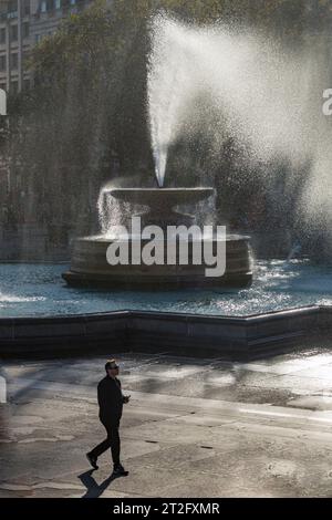 Uomo che cammina davanti a una delle fontane di Trafalgar Square, Londra, in una ventosa giornata autunnale Foto Stock