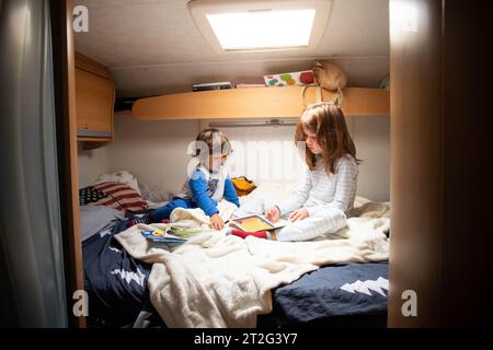 Due bambini caucasici, fratello e sorella, leggono libri in pigiama su un letto di camper durante una sosta in viaggio. Vacanze in camper con bambini Foto Stock