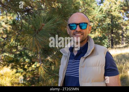 Ritratto sorridente 40 anni uomo con occhiali da sole, albero di abete, foresta sullo sfondo. La natura in autunno lascia la stagione. Piano orizzontale. Stile di vita attivo. Maschio Foto Stock