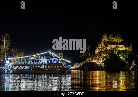 Barca illuminata ancorata nella tranquilla baia con piccola isola nella baia di Parga, Grecia, di notte. Le acque calme del mare riflettono le luci. Foto Stock