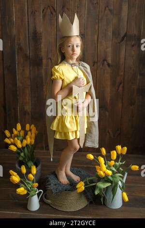 Ritratto di una principessa ragazza in una corona di cartone con una spada in mano, accanto a mazzi di tulipani gialli. Telaio verticale. Guarda la telecamera. Foto Stock