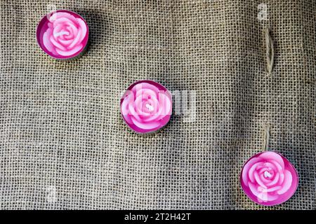 Tre bellissime candele in cera rosa sotto forma di fiori di rosa con uno stoppino non cotto sullo sfondo di una vecchia tela marrone, un panno duro e non sbiancato ma Foto Stock