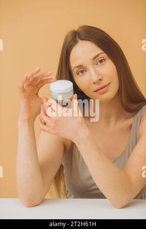 Ritratto alla moda di una bella donna con una bottiglia di crema cosmetica su sfondo pastello beige. Foto Stock