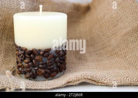 Cera bella candela beige chiaro con stoppino non aromatizzato dal basso decorata con chicchi di caffè sullo sfondo di un vecchio tessuto di tela marrone. Foto Stock