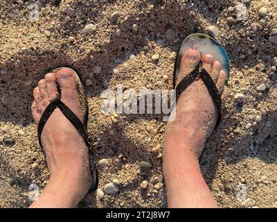 Primo piano di un uomo con pantofole nere in piedi sulla spiaggia, con un'onda di schiuma delicata sotto di loro. Vista dall'alto. Foto Stock