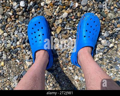 Gambe da uomo, piedi in pantofole di gomma blu sullo sfondo di ciottoli e sabbia in una vacanza sulla spiaggia in un caldo paradiso tropicale orientale Foto Stock
