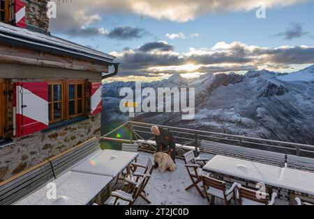 Uomo con cane di fronte all'alba sopra il rifugio Ramolhaus con vista sulla cresta principale ricoperta di nubi delle Alpi Ötztal e Stubai Foto Stock