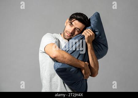 Bellissimo maschio barbato caucasico in t-shirt che abbraccia un morbido cuscino mentre si trova in piedi su uno sfondo grigio isolato in studio Foto Stock