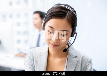 Donna asiatica concentrata in abiti formali e cuffia con microfono che lavora in call center con un collega su sfondo sfocato Foto Stock