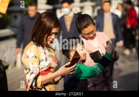 Una donna vietnamita vestita in abito tradizionale ao dai guarda il suo telefono cellulare con un amico e un ragazzo alla Pagoda Tran Quoc di Hanoi, in Vietnam, a. Foto Stock