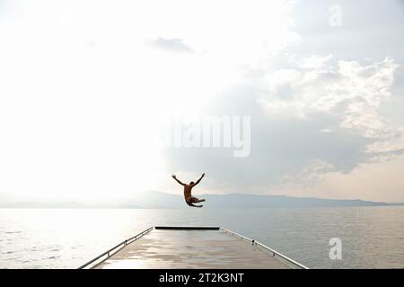 Un giovane adolescente tira le braccia e calza le gambe in aria mentre salta da un molo per una nuotata nell'acqua fresca del lago Flathead, Montana. Foto Stock