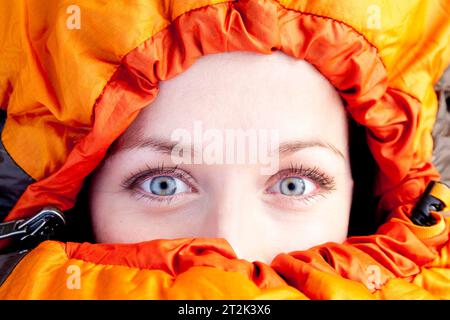 Una donna dagli occhi blu brillante guarda direttamente nella fotocamera mentre è stretta nel sacco a pelo arancione. Foto Stock