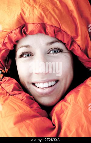 Una donna dagli occhi blu brillante guarda direttamente nella fotocamera mentre è stretta nel sacco a pelo arancione. Foto Stock