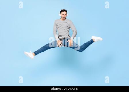 Giovane uomo caucasico divertente ed energico che indossa cuffie che ascolta musica e salta con gambe spalancate in uno studio di colore blu chiaro su sfondo isolato Foto Stock