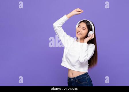 Allegra, felice e sorridente donna asiatica che ascolta musica con le cuffie e balla in uno studio di colore viola con sfondo isolato Foto Stock