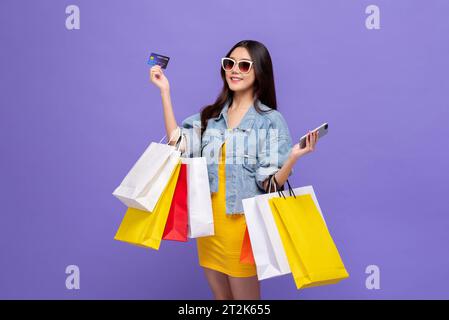 Bellissima giovane donna asiatica sorridente che porta borse colorate e mostra la carta di credito in uno studio isolato, concetto di shopping online mobile Foto Stock