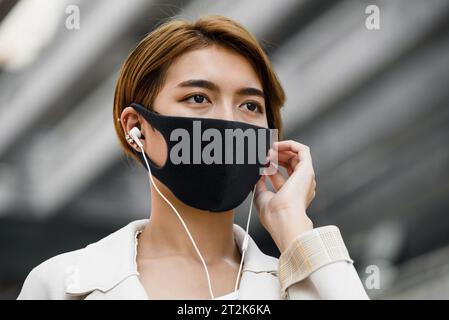 Giovane donna asiatica che indossa la maschera facciale all'aperto mentre ascolta musica in città durante la pandemia di COVID-19 Foto Stock